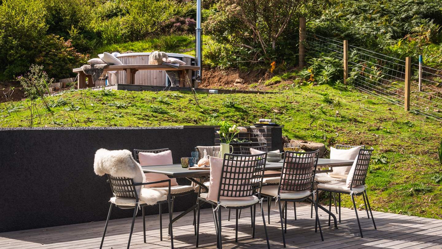 Calling for sun-kissed moments and peaceful evenings under wide skies, a large wooden deck resides outside with a dining table and cushion-topped chairs for eight people and a built-in BBQ